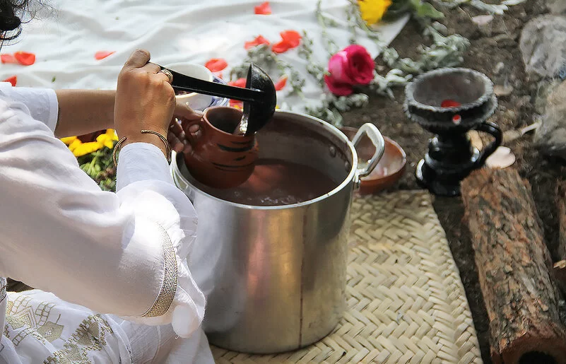 Eine Frau schöpft mit einer Kelle eine Flüssigkeit aus einem Topf in eine Tasse. Im Hintergrund sieht man Blumen.