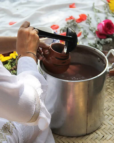 Eine Frau schöpft mit einer Kelle eine Flüssigkeit aus einem Topf in eine Tasse. Im Hintergrund sieht man Blumen.