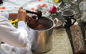 Eine Frau schöpft mit einer Kelle eine Flüssigkeit aus einem Topf in eine Tasse. Im Hintergrund sieht man Blumen.