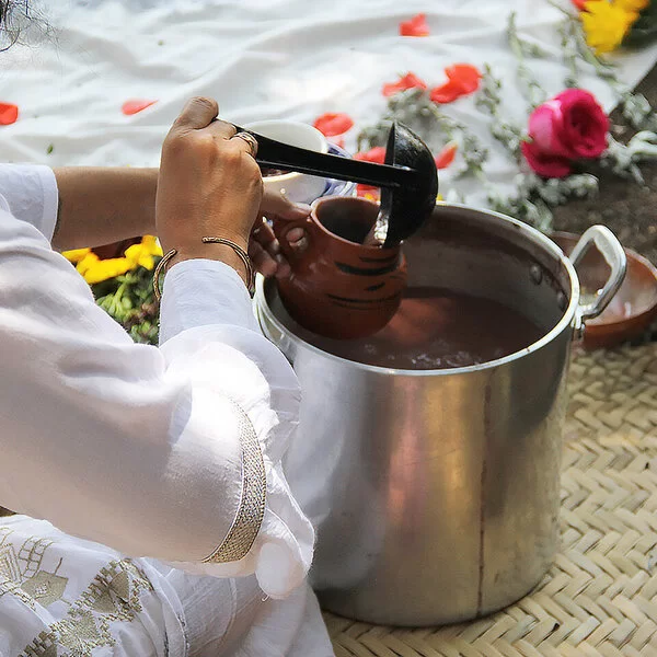 Eine Frau schöpft mit einer Kelle eine Flüssigkeit aus einem Topf in eine Tasse. Im Hintergrund sieht man Blumen.