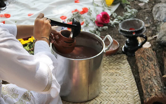 Eine Frau schöpft mit einer Kelle eine Flüssigkeit aus einem Topf in eine Tasse. Im Hintergrund sieht man Blumen.