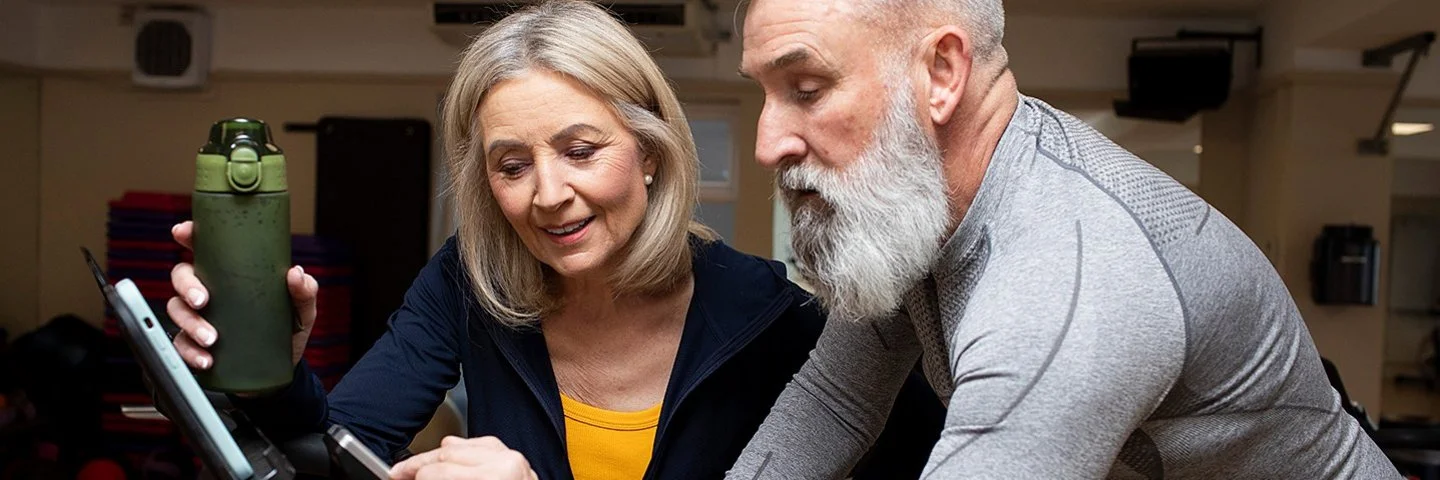 Ein älterer Mann mit weißem Vollbart sitzt in grauer Sportkleidung auf einem Trainingsfahrrad im Fitnessstudio. Er zeigt einer älteren Frau neben ihm etwas auf dem Display des Geräts. Die Frau hält eine grüne Trinkflasche in der Hand und trägt eine dunkelblaue Trainingsjacke. Beide wirken konzentriert und freundlich.