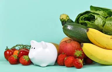 Ein kleines Sparschwein ist von gesundem Obst und Gemüse umgeben.