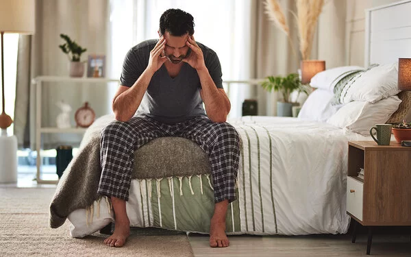 Ein mittelalter Mann mit dunklen Haaren sitzt auf einem Bett und hält mit beiden Händen seinen nach vorn gebeugten Kopf, während er die Arme auf den Oberschenkeln abstützt.