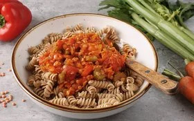 Weißer Teller mit Vollkorn-Fusilli und Gemüsebolognese, darin steckt ein Löffel mit Holzgriff. Daneben und dahinter liegen auf grauem Hintergrund einige rote Linsen, rote Paprika, Möhren und Stangensellerie.