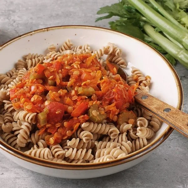 Weißer Teller mit Vollkorn-Fusilli und Gemüsebolognese, darin steckt ein Löffel mit Holzgriff. Daneben und dahinter liegen auf grauem Hintergrund einige rote Linsen, rote Paprika, Möhren und Stangensellerie.