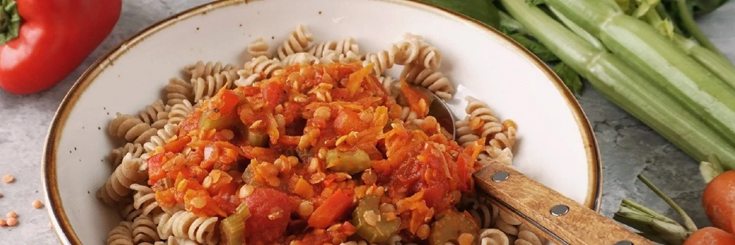 Weißer Teller mit Vollkorn-Fusilli und Gemüsebolognese, darin steckt ein Löffel mit Holzgriff. Daneben und dahinter liegen auf grauem Hintergrund einige rote Linsen, rote Paprika, Möhren und Stangensellerie.