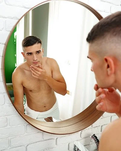 Ein junger Mann mustert sein Gesicht kritisch im Badezimmerspiegel.
