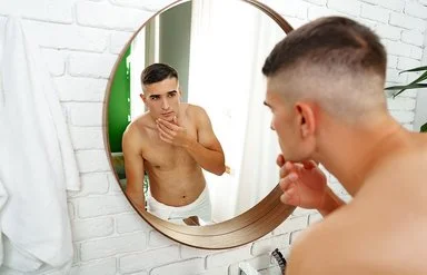 Ein junger Mann mustert sein Gesicht kritisch im Badezimmerspiegel.