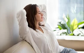 Eine Frau sitzt mit geschlossenen Augen und mit den Armen hinter dem Kopf verschränkt auf dem Sofa und atmet tief durch.