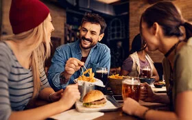 Ein Mann und zwei Frauen essen Hamburger und Pommes frites in einem Restaurant und trinken Bier dazu.