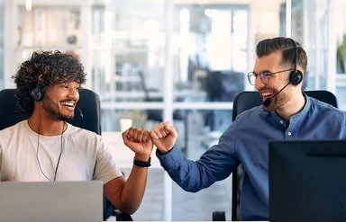 Zwei Männer sitzen nebeneinander an einem Tisch, vor ihnen stehen Computerbildschirme. Der linke Mann hat kurzes gelocktes Haar und einen Vollbart. Er trägt ein weißes T-Shirt und an der linken Hand eine Uhr. Der rechte Mann trägt ein blaues Hemd. Er hat kurze Haare, einen Dreitagebart und trägt eine Brille. Beide Männer haben ein Headset um. Sie lächeln sich an und drücken ihre Fäuste zur Siegerpose aneinander.
