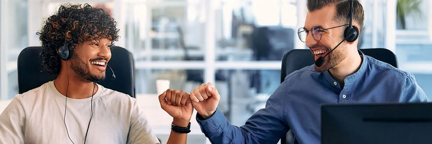 Zwei Männer sitzen nebeneinander an einem Tisch, vor ihnen stehen Computerbildschirme. Der linke Mann hat kurzes gelocktes Haar und einen Vollbart. Er trägt ein weißes T-Shirt und an der linken Hand eine Uhr. Der rechte Mann trägt ein blaues Hemd. Er hat kurze Haare, einen Dreitagebart und trägt eine Brille. Beide Männer haben ein Headset um. Sie lächeln sich an und drücken ihre Fäuste zur Siegerpose aneinander.
