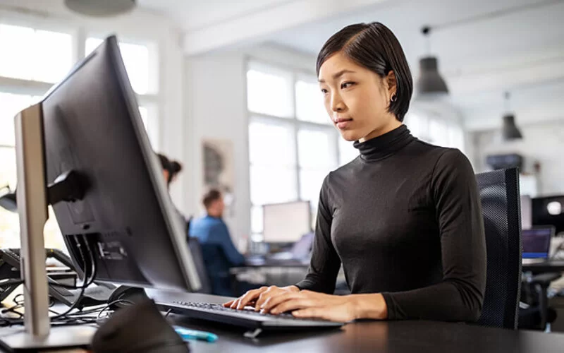 Eine Frau sitzt in einem Großraumbüro an ihrem Schreibtisch und arbeitet an ihrem Computer.