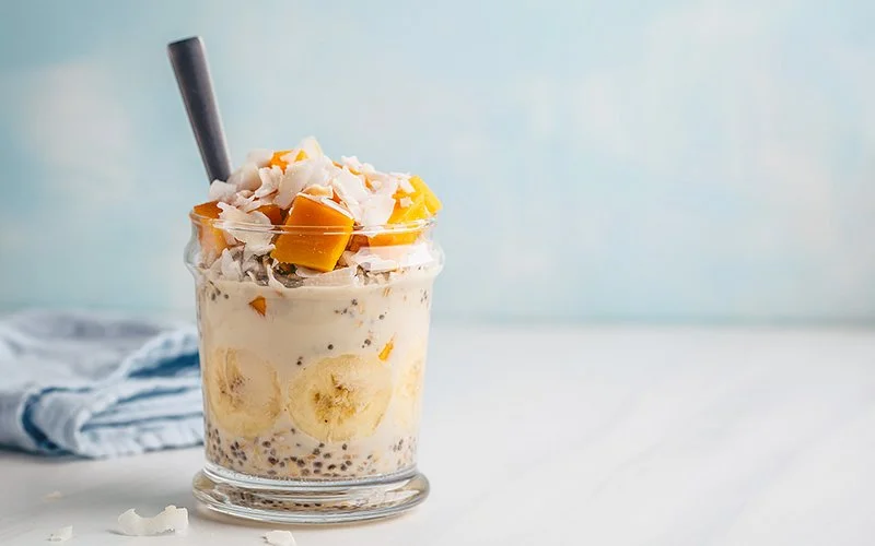 In einem Glas sind Overnight Oats mit Bananenscheiben, Mangostückchen, Chia-Samen und Kokosflocken verfeinert.