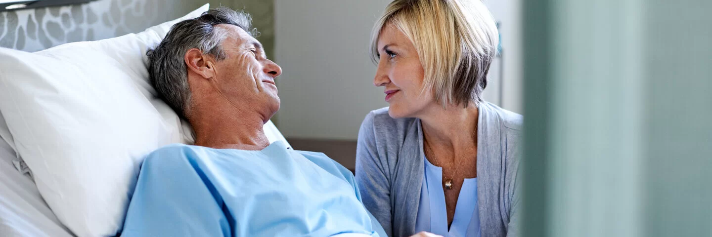 Ein Mann liegt im Krankenhaus; seine Frau spricht mit ihm. Vor schweren Eingriffen sollte man eine Patientenverfügung verfassen.