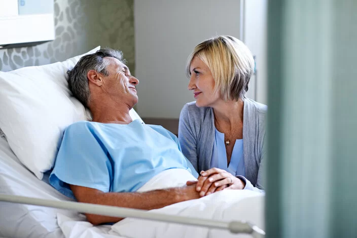 Ein Mann liegt im Krankenhaus; seine Frau spricht mit ihm. Vor schweren Eingriffen sollte man eine Patientenverfügung verfassen.