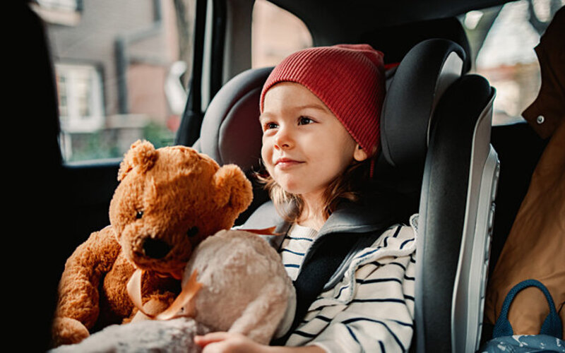 Ein kleines Mädchen spielt mit einem Teddy während sie in der Sitzerhöhung im Auto sitzt.