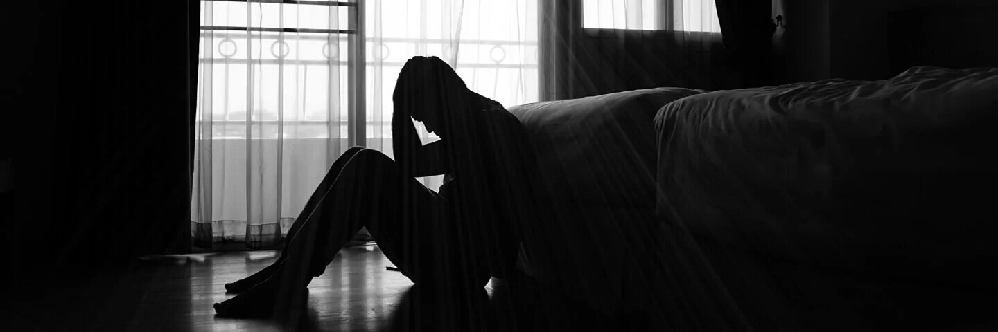 Ein Mann mit Suizidgedanken sitzt in einem dunklem Zimmer vor dem Bett.
