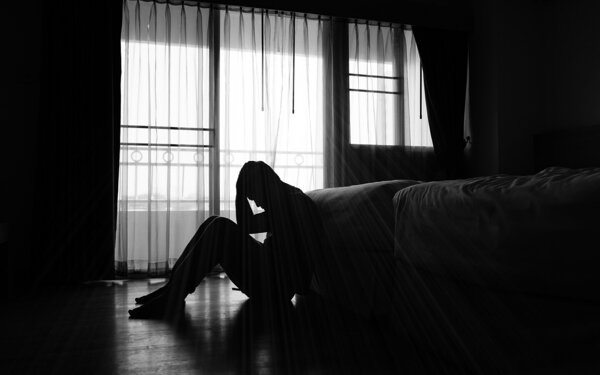 Ein Mann mit Suizidgedanken sitzt in einem dunklem Zimmer vor dem Bett.