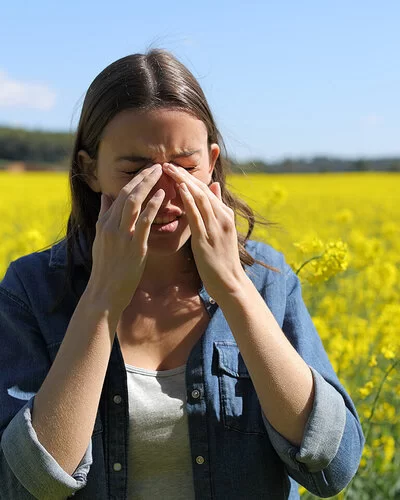Eine Frau steht in einem Rapsfeld und fast sich mit beiden Händen an die Nase. Bei Pollenallergie hilft es, ein Allergie-Tagebuch zu führen.
