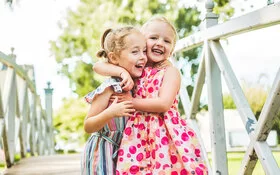 Zwei etwa dreijährige Mädchen in bunten Kleidern in einem Park auf einer Holzbrücke. Sie lachen und halten sich fest in den Armen.
