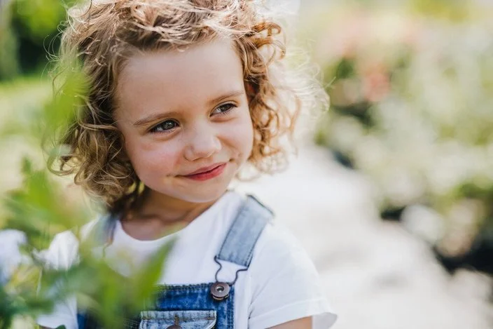 Porträt eines kleinen Mädchens mit Locken und Latzhose im Garten.