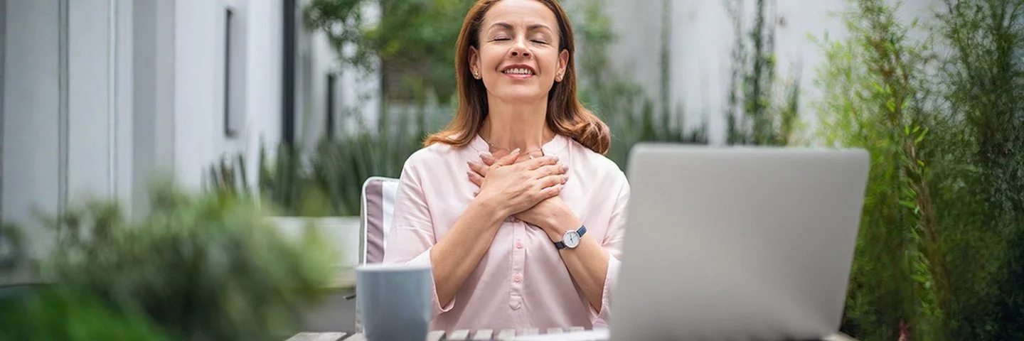 Eine Frau sitzt an einem Holztisch im Freien vor einem Laptop mit einer Tasse und hält beide Hände an die Brust