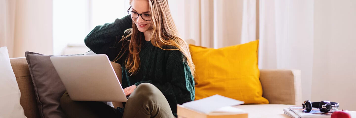 Eine junge blonde Frau mit Brille sitzt auf einem Sofa und schaut auf einen Laptop.