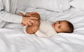 Eine Mutter macht Hüft-Gymnastik mit ihrem Baby, das auf dem Bett liegt.