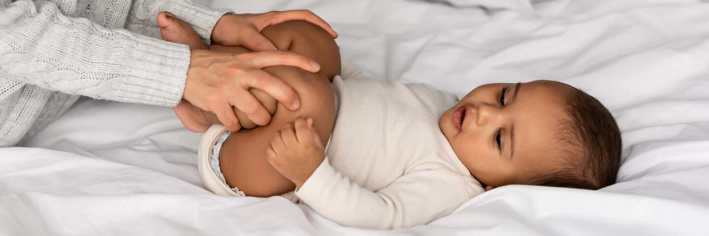 Eine Mutter macht Hüft-Gymnastik mit ihrem Baby, das auf dem Bett liegt.