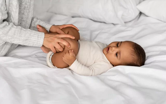 Eine Mutter macht Hüft-Gymnastik mit ihrem Baby, das auf dem Bett liegt.