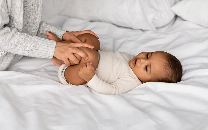 Eine Mutter macht Hüft-Gymnastik mit ihrem Baby, das auf dem Bett liegt.