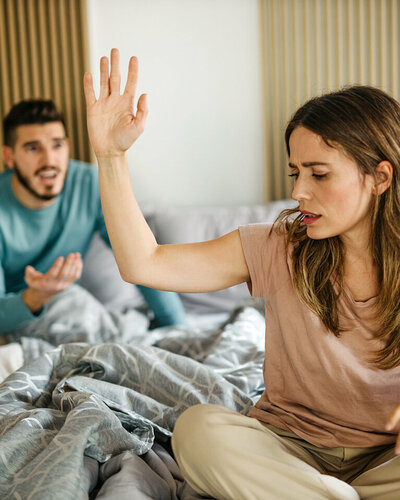 Ein junges Paar sitzt streitend auf dem Bett, sie wehrt mit einer Handbewegung seine Äußerungen ab.