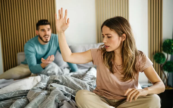 Ein junges Paar sitzt streitend auf dem Bett, sie wehrt mit einer Handbewegung seine Äußerungen ab.