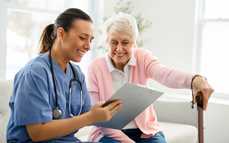 Eine weibliche Pflegefachperson besucht eine ältere Frau zu Hause und zeigt ihr etwas auf dem Tablet.