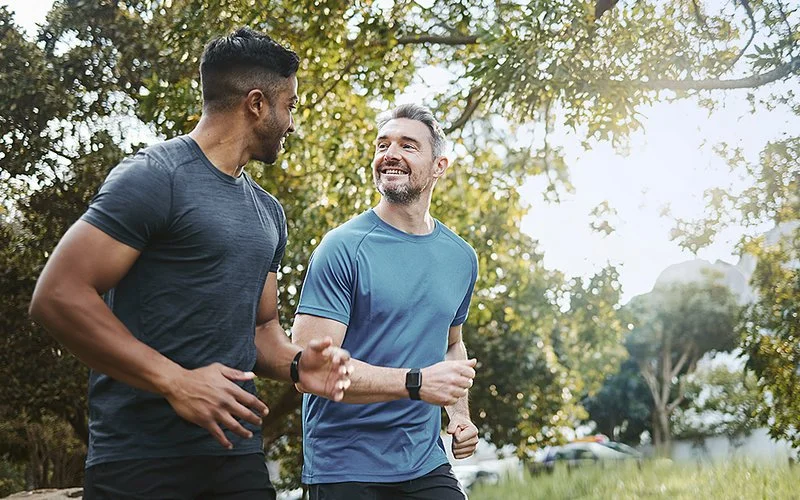Zwei Männer joggen durch einen Naturpark. Sie schauen sich gegenseitig an und lächeln.