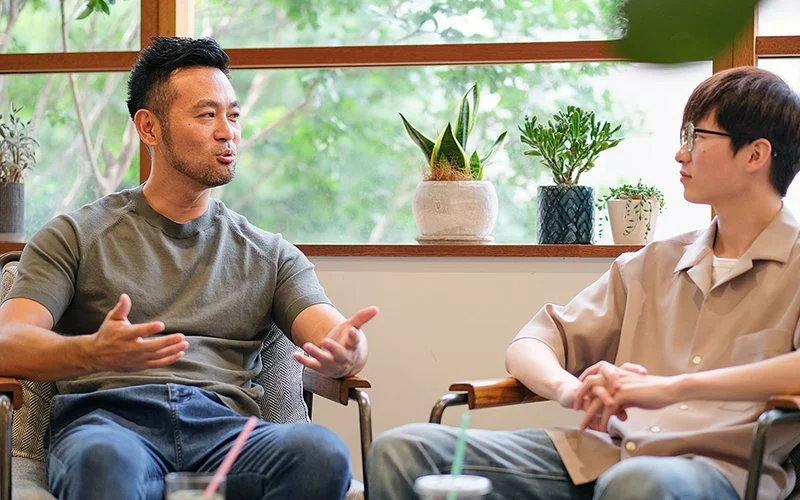 In einem hellen Raum mit großen Fenstern und Zimmerpflanzen im Hintergrund sitzen sich zwei Männer in Sesseln gegenüber und führen ein Gespräch.