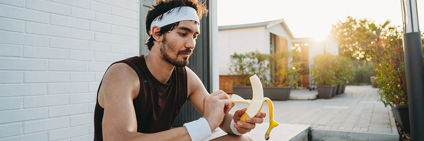 Ein junger Mann sitzt in schwarzer Sportkleidung auf einer Treppenstufe und schält eine Banane.