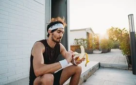 Ein junger Mann sitzt in schwarzer Sportkleidung auf einer Treppenstufe und schält eine Banane.