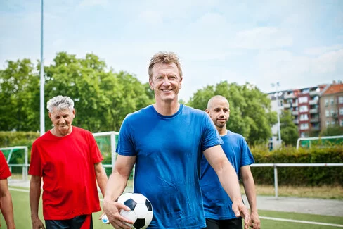Drei Männer stehen auf einem Fußballplatz, einer von ihnen hält einen Fußball in der Hand.
