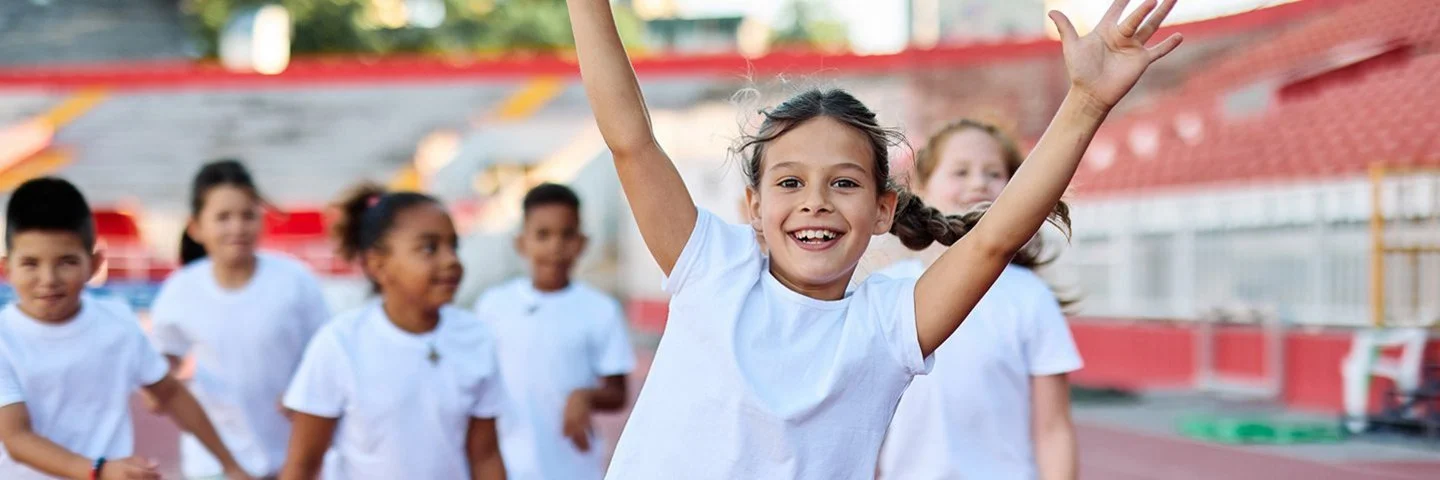 Gruppe fröhlicher Kinder in Sportkleidung läuft auf einer Laufbahn, vorne ein Mädchen mit erhobenen Armen