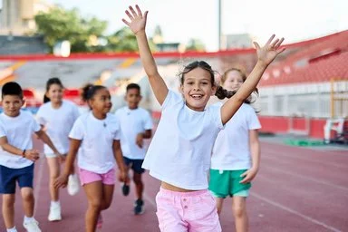 Gruppe fröhlicher Kinder in Sportkleidung läuft auf einer Laufbahn, vorne ein Mädchen mit erhobenen Armen