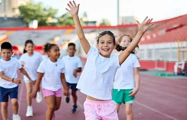 Gruppe fröhlicher Kinder in Sportkleidung läuft auf einer Laufbahn, vorne ein Mädchen mit erhobenen Armen