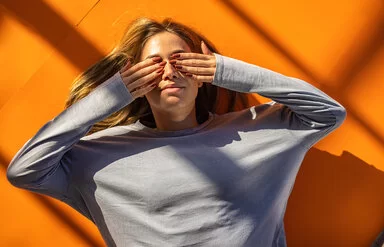 Eine junge Frau liegt rücklings auf orangefarbenem Boden und hält sich mit den Händen die Augen zu.