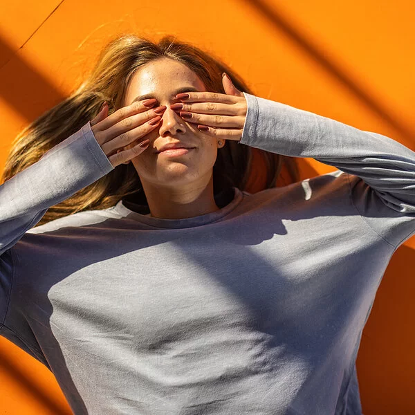 Eine junge Frau liegt rücklings auf orangefarbenem Boden und hält sich mit den Händen die Augen zu.