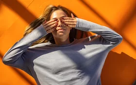 Eine junge Frau liegt rücklings auf orangefarbenem Boden und hält sich mit den Händen die Augen zu.