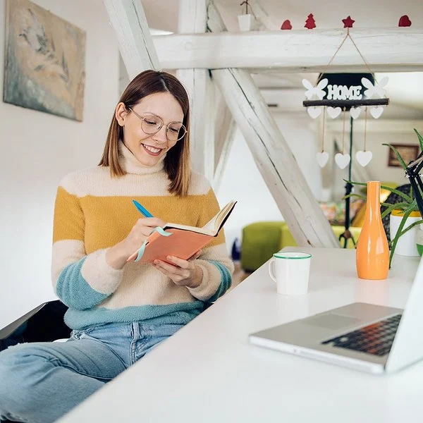 Eine Frau sitzt entspannt an einem Schreibtisch in einem hellen, weihnachtlich geschmückten Zimmer. Sie hält ein Notizbuch in der Hand und schreibt lächelnd hinein. Vor ihr stehen ein Laptop und eine Tasse.