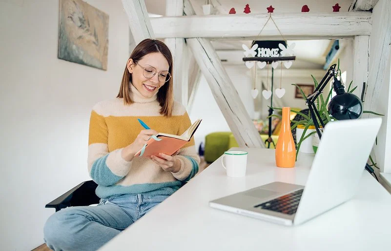 Eine Frau sitzt entspannt an einem Schreibtisch in einem hellen, weihnachtlich geschmückten Zimmer. Sie hält ein Notizbuch in der Hand und schreibt lächelnd hinein. Vor ihr stehen ein Laptop und eine Tasse.