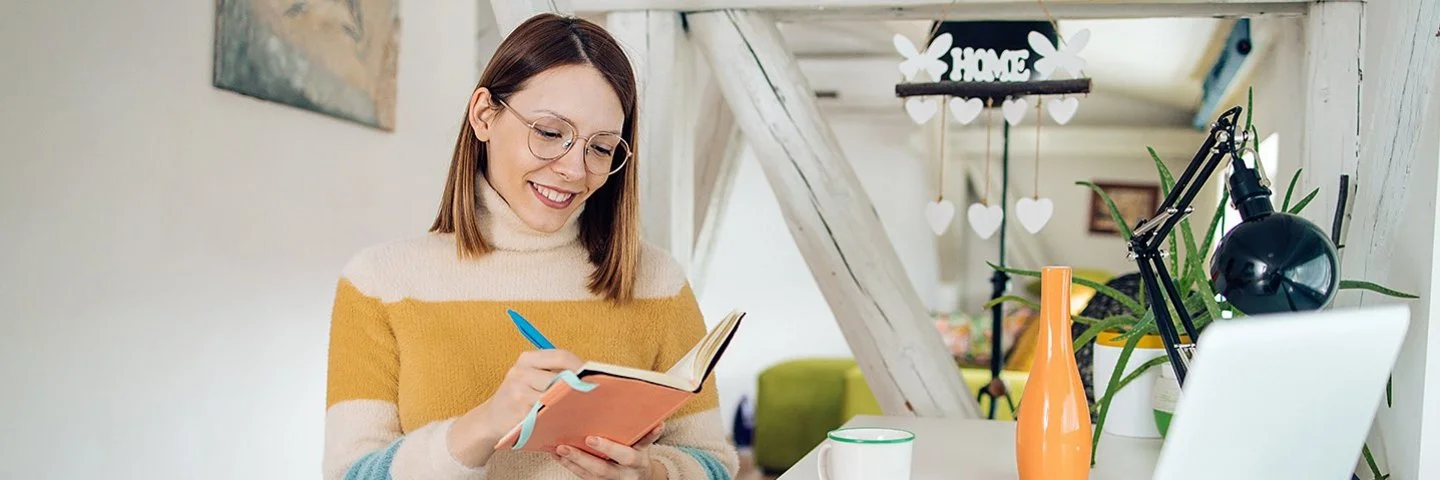 Eine Frau sitzt entspannt an einem Schreibtisch in einem hellen, weihnachtlich geschmückten Zimmer. Sie hält ein Notizbuch in der Hand und schreibt lächelnd hinein. Vor ihr stehen ein Laptop und eine Tasse.